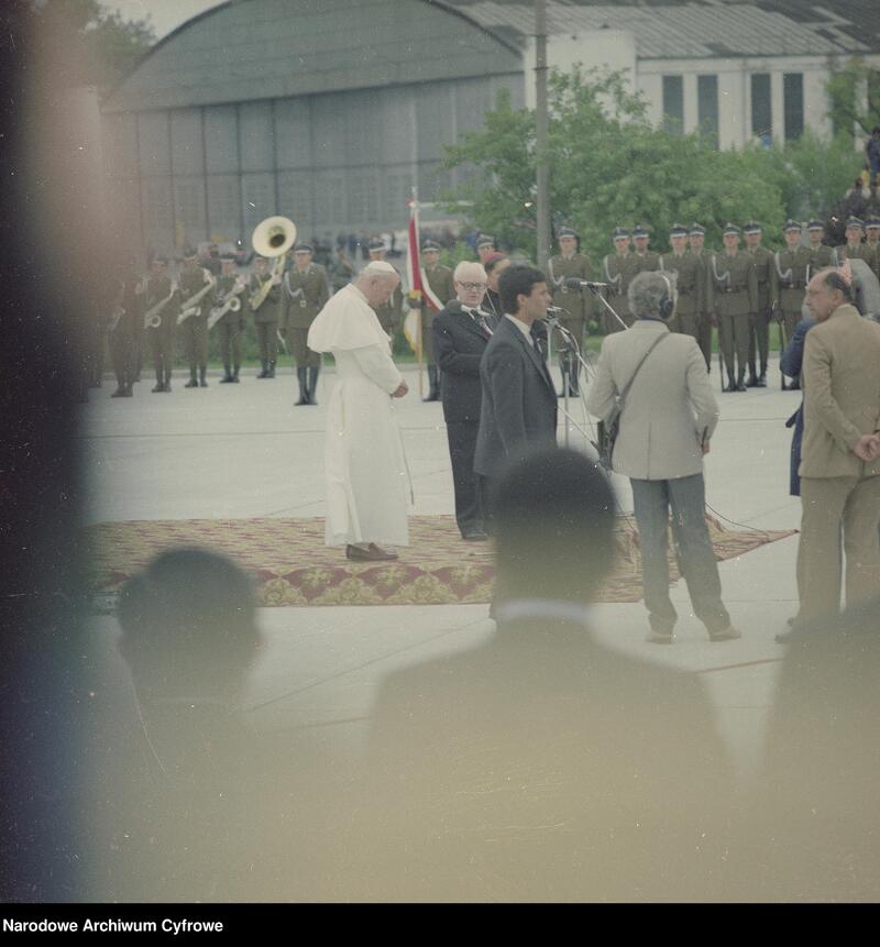 Na podium z lewej widoczny Jan Paweł II. Z prawej przed podium widoczni fotoreporterzy, w głębi żołnierze z Kampanii Reprezentacyjnej Wojska Polskiego, w tym orkiestra i poczet sztandarowy.