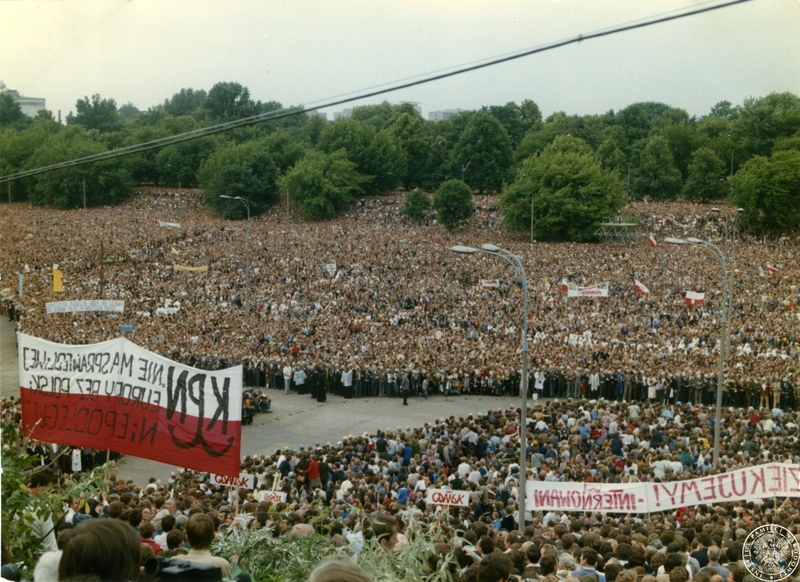 Fot. nr 1: Tłum wiernych na błoniach przed Stadionem X-lecia oczekuje na przyjazd Jana Pawła II. Na pierwszym planie po lewej biało-czerwony transparent, widziany od lewej strony: KPN. NIE MA / SPRAWIEDLIWEJ EUROPY BEZ POLSKI / NIEPODLEGŁEJ, pośrodku małe transparenty z napisami: GDAŃSK, w prawym dolnym rogu biały transparent z czerwonym napisem: [KOCHAMY CIĘ!] DZIĘKUJEMY! - INTERNOWANI. W górnej części po lewej stronie biały transparent z niebieskim napisem: CHRYSTUS NIE ZGODZI SIĘ NIGDY ABY CZŁOWIEK / ZOSTAŁ UZNANY TYLKO ZA NARZĘDZIE PRACY, po prawej biały transparent z napisem: OJCZE ŚWIĘTY WITAMY / INTERNOWANI. 17 VI 1983 r. Sygnatura: IPNBU-7-14-2-1
