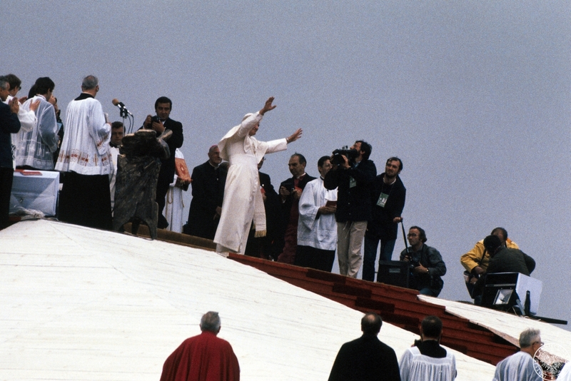 Fot. nr 8. Jan Paweł II otoczony przez duchownych i fotoreporterów pozdrawia wiernych z podwyższenia ołtarza w czasie nabożeństwa na lotnisku w dzielnicy Muchowiec. 20 VI 1983 r. Sygnatura: IPNBU-7-14-13-76